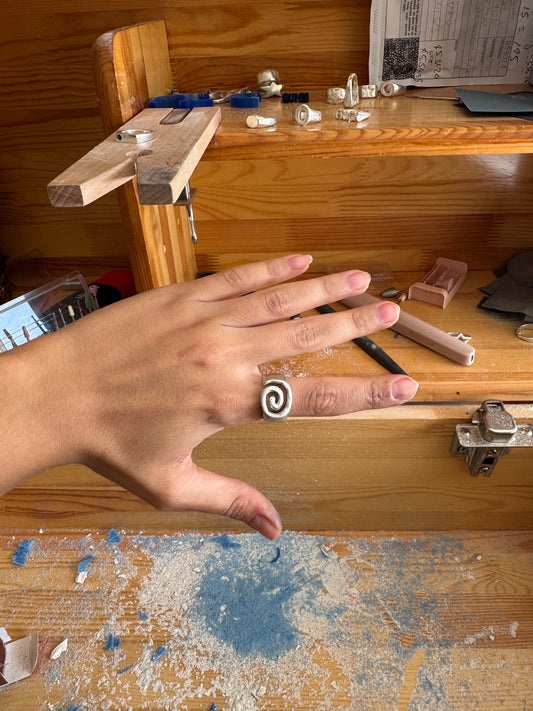 Hand with a silver swirl ring on a wooden workbench with tools and materials